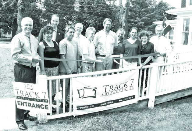 Pictured here at the grand opening of the Track 7 Postal Center's new location are Orange County Legislator Barry Cheney (left) and Mayor Michael Newhard (right) holding the ribbon as Betsy and Ken Mitchel, owners of Track 7l (center) prepare to cut it to celebrate the grand opening. They are surrounded by family and friends.