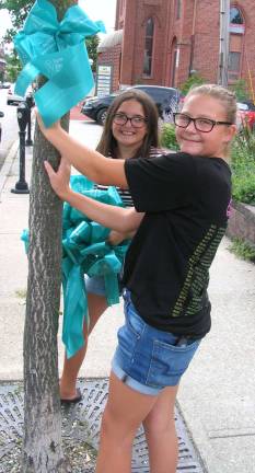 Middle School students Kailyn Ackerman, 11 (left), and Emma Kardell, 12, place one of the teal-colored ribbons on a tree along Main Street.