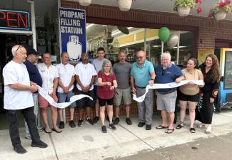 Jane Fink cuts the anniversary ribbon, as Bernie Fink (far left in white) and Mayor Howley (in turquoise shirt), Village Trustee Matt Veth (in navy shirt), Greenwood Lake Chamber President, Brenda Wiley (far right), and staff and community members look on.