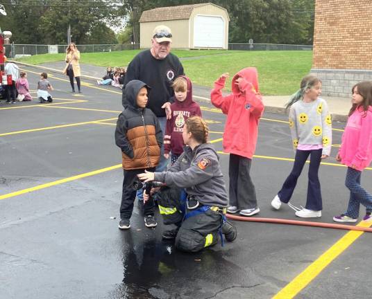 Firefighter Kaitlyn Wendel shows a student at Park Avenue Elementary School how to use a nozzle while Safety Officer Michael Sinclair assists.