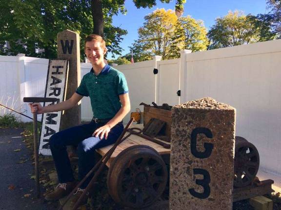 Alex Prizgintas is seated on his own growing railroad, a tool cart which stands between a concrete whistle post (left) and a mile marker from Craigville, N.Y. (right) in his backyard. His newest acquisition can be seen leaning against the whistle post &#x2014; the Harriman station sign which he was able to get from a dealer from Ontario, Canada.